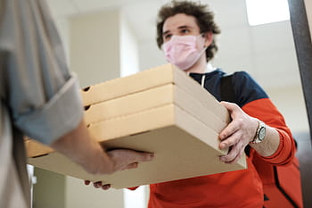 Delivery worker handing over pizzas while wearing a protective mask indoors, illustrating safe delivery practices during a pandemic. HD wallpaper