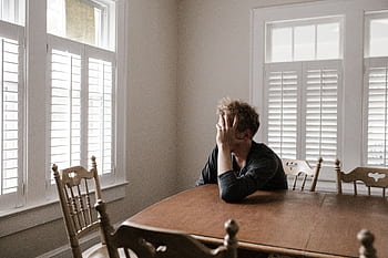 A man sits alone at a table in a bright room, displaying deep contemplation. HD wallpaper
