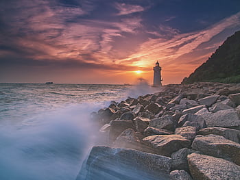 The sun sets beautifully behind a seaside lighthouse, creating a dramatic landscape view. HD wallpaper