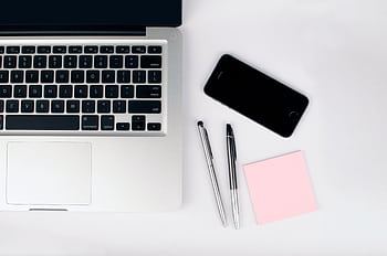 A top view of a modern workspace featuring a laptop, smart, pens, and a pink sticky note on a white desk. HD wallpaper
