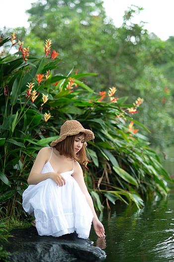 A serene portrait of a young woman in a white dress sitting by a stream, surrounded by lush greenery. HD phone wallpaper