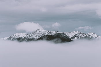 A serene winter view of snow-capped mountains rising above misty clouds on a cloudy day. HD wallpaper