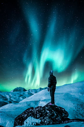 A lone figure stands on a snowy slope, gazing at the vibrant Northern Lights under a starry sky. HD phone wallpaper