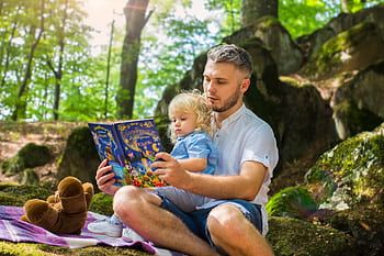 Father and toddler bonding over a book in a sunny forest setting. HD wallpaper