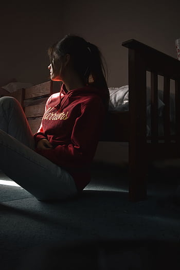 Contemplative young woman in hoodie sitting on the floor in dimly lit room, symbolizing solitude and introspection. HD phone wallpaper