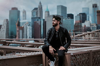 A man wearing a black leather jacket sitting on Brooklyn Bridge with New York City skyline in the background. HD wallpaper