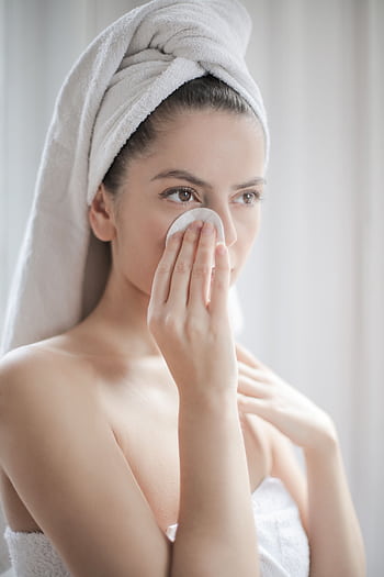 A woman using a cotton pad for skincare, wrapped in a towel indoors. HD phone wallpaper