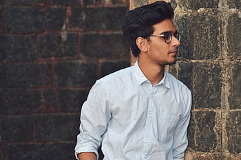 A stylish young man in glasses leans against a brick wall wearing a casual light blue shirt. HD wallpaper