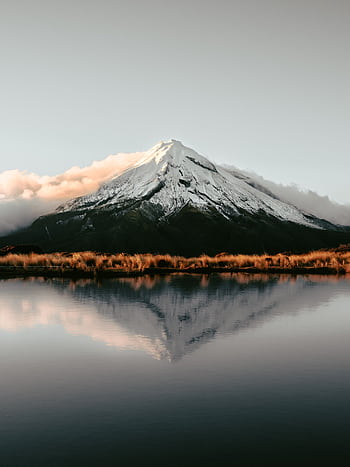 Tranquil view of snow-capped Mount Taranaki reflecting in a serene lake under soft, warm light. HD phone wallpaper