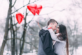 A loving couple embracing outdoors with heart-shaped balloons during a snowy winter day. HD wallpaper