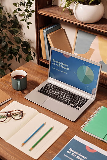 Laptop displaying ad spend data on a wooden desk with office supplies. HD phone wallpaper