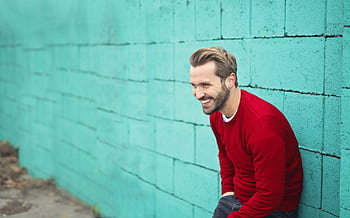 Smiling man in Budapest wearing a red sweater against a teal brick wall, exuding happiness. HD wallpaper