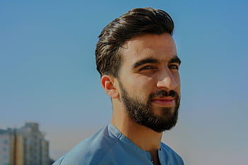 A close-up outdoor portrait of a young man with facial hair, exuding confidence under a clear sky. HD wallpaper