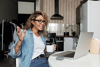 Smiling woman with glasses having a video call from home, enjoying a cup of coffee in her kitchen. HD wallpaper