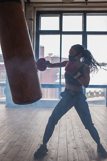 A woman boxer in a mask trains with a punching bag indoors, exemplifying focus and strength. HD phone wallpaper