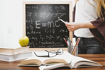 Woman writing physics equations on a blackboard with books and an apple on the desk. HD wallpaper