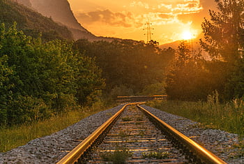 Golden sunset over railway tracks surrounded by lush greenery and mountains. HD wallpaper