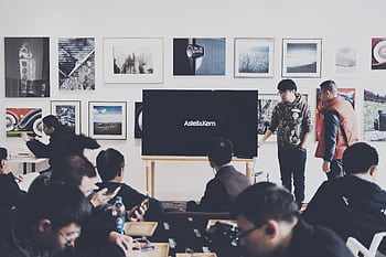 A group of adults attending a casual business presentation in a modern art space, featuring a TV display and framed art. HD wallpaper