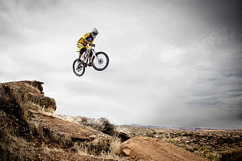 A cyclist performs a daring jump off a rocky cliff under a cloudy sky. HD wallpaper