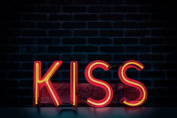 Bright neon lit 'Kiss' sign with a dark brick wall background, ideal for modern decor themes. HD wallpaper