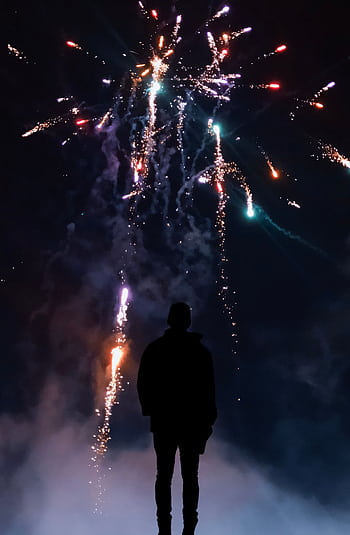 A person silhouetted against a colorful night sky with fireworks bursting overhead. HD phone wallpaper