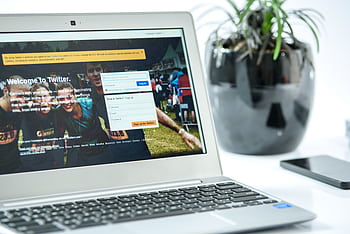 Close-up of a laptop displaying Twitter login page, accompanied by a smart and a potted plant in a bright workspace. HD wallpaper