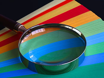 Magnifying glass resting on vibrant rainbow-colored paper close-up. HD wallpaper