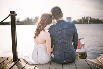Bride and groom sitting on a dock by a serene lake, enjoying a romantic moment under the sun. HD wallpaper