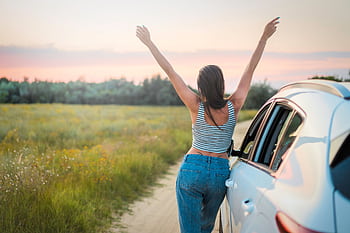 A woman stands beside a car on a summer field road, arms raised in dom, enjoying a beautiful sunset. HD wallpaper
