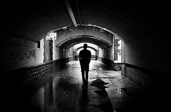 A person walking alone through a dimly lit tunnel, captured in black and white. HD wallpaper