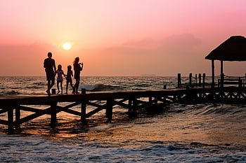 Silhouetted family enjoys a stroll on the beach pier at a vibrant sunset over the ocean waves. HD wallpaper