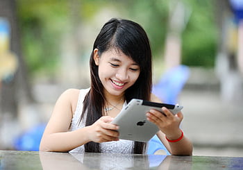 Smiling young woman using a tablet while sitting outdoors, enjoying leisure time in a park during a sunny day. HD wallpaper