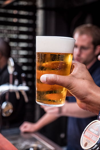 Close-up of a hand holding a foamy beer glass with blurred bar background. HD phone wallpaper