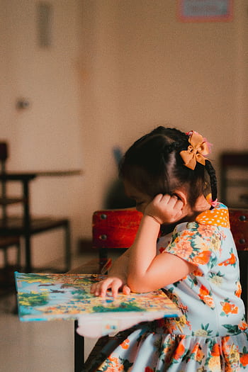 A young girl engrossed in reading a book in an indoor classroom setting. HD phone wallpaper