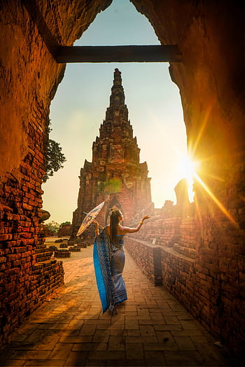 A woman in traditional wear stands with an umbrella at a historic Ayutthaya temple during sunset. HD phone wallpaper