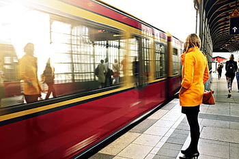 A woman in a yellow coat waits as a red train passes in a bustling station. HD wallpaper