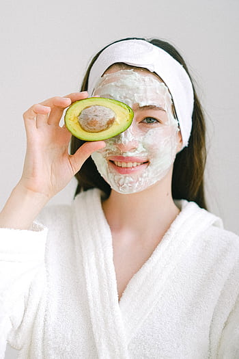 Smiling woman with face mask holding avocado for skincare in spa setting. HD phone wallpaper