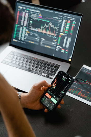 Person analyzing stock market data on a smart with a laptop displaying financial graphs in the background. HD phone wallpaper