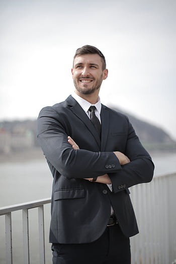 Smiling businessman in a suit standing confidently outdoors by the river. HD phone wallpaper