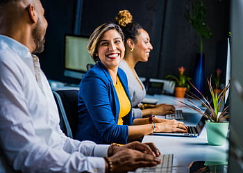 Three diverse professionals working and smiling at office desks, fostering teamwork and collaboration. HD wallpaper