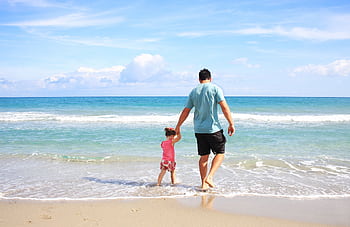 A father and daughter hold hands while walking on a sunny beach, enjoying family time together. HD wallpaper