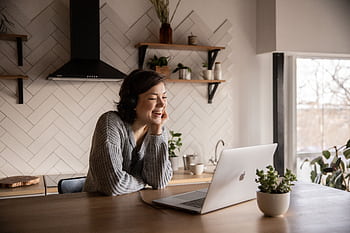 Smiling woman in a cozy kitchen engaged in a joyful video call using her laptop. HD wallpaper