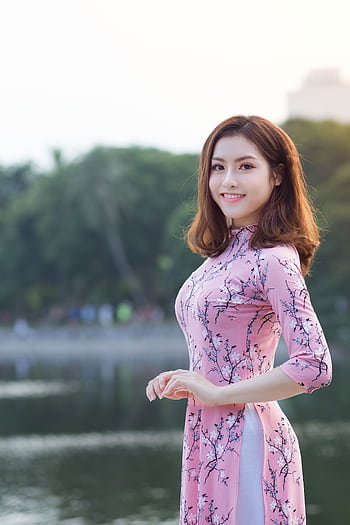 Asian woman in a pink floral dress smiling by a serene lake, surrounded by nature. HD phone wallpaper