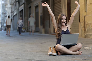 A cheerful woman sitting outdoors, celebrating success with arms raised, while using a laptop. HD wallpaper