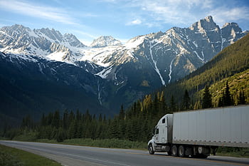 A semi-truck travels along a highway with snow-capped mountains in the background. HD wallpaper