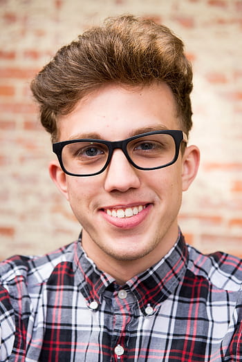 Portrait of a smiling young man wearing eyeglasses and plaid shirt against a brick wall background. HD phone wallpaper