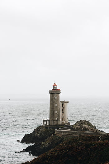 A solitary lighthouse stands on a rocky shore, overlooking a misty and tranquil ocean. HD phone wallpaper
