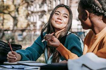 Two diverse female students studying together outside with notebooks and pens, expressing joy and collaboration. HD wallpaper