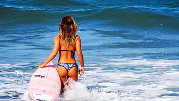 A young woman enters the ocean with her pink surfboard, ready to surf the waves on a sunny day. HD wallpaper