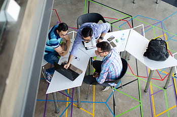 Three men collaborating over a laptop in a modern, geometric-themed office space. HD wallpaper